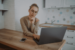 Beautiful smiling girl in glasses in knitted sweater using laptop at kitchen