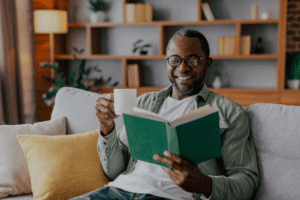 Cheerful adult african american man in glasses and casual reading book and enjoy favorite drink on